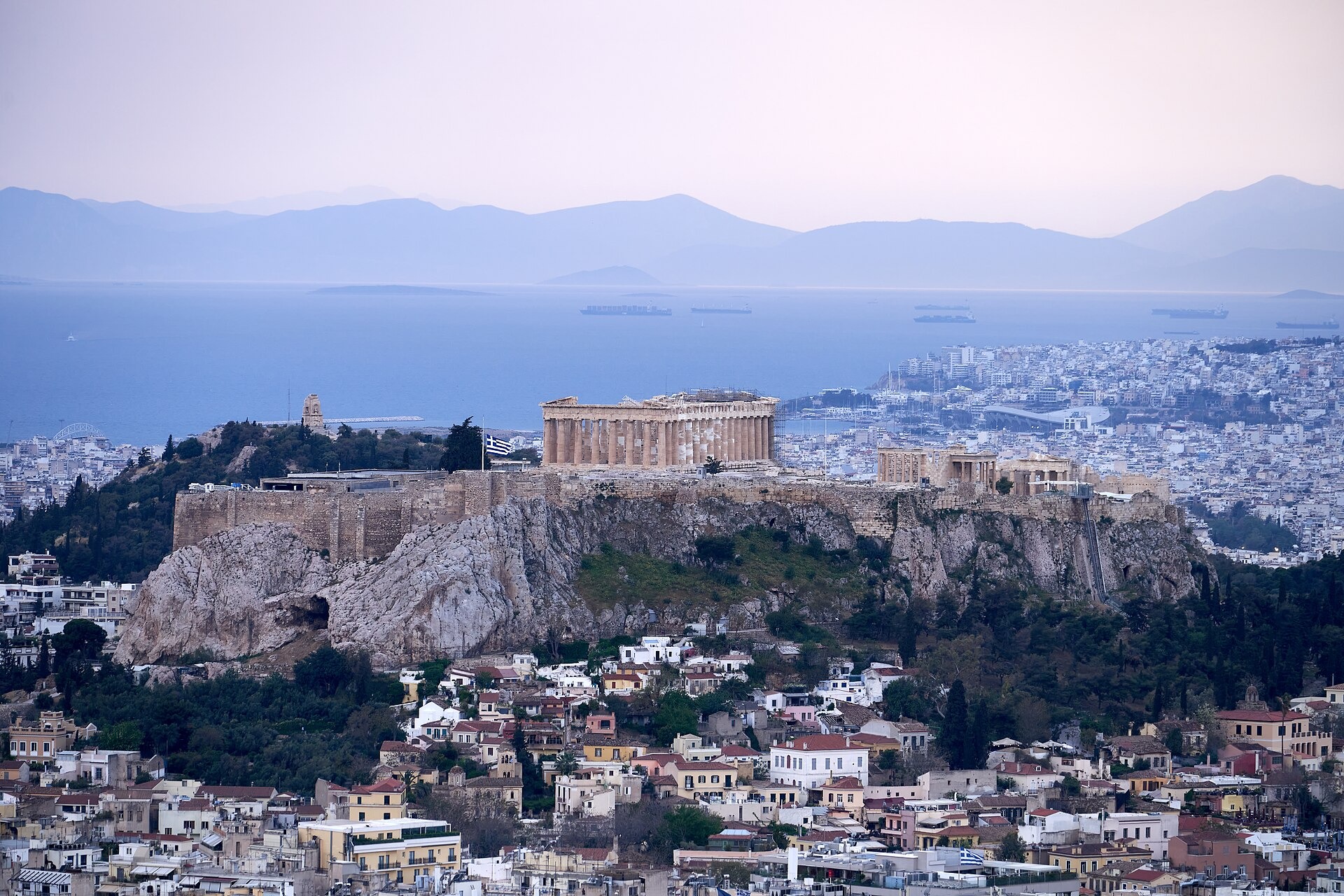 The Acropolis of Athens as seen from Mount Lycabettus The wooded Hill of the Nymphs is half-visible on its right, and Philopappos Hill on the left, immediately behind. The Philopappos Monument is seen here where, in the distant background, the coast of Peloponnese meets the waters of the Saronic Gulf.