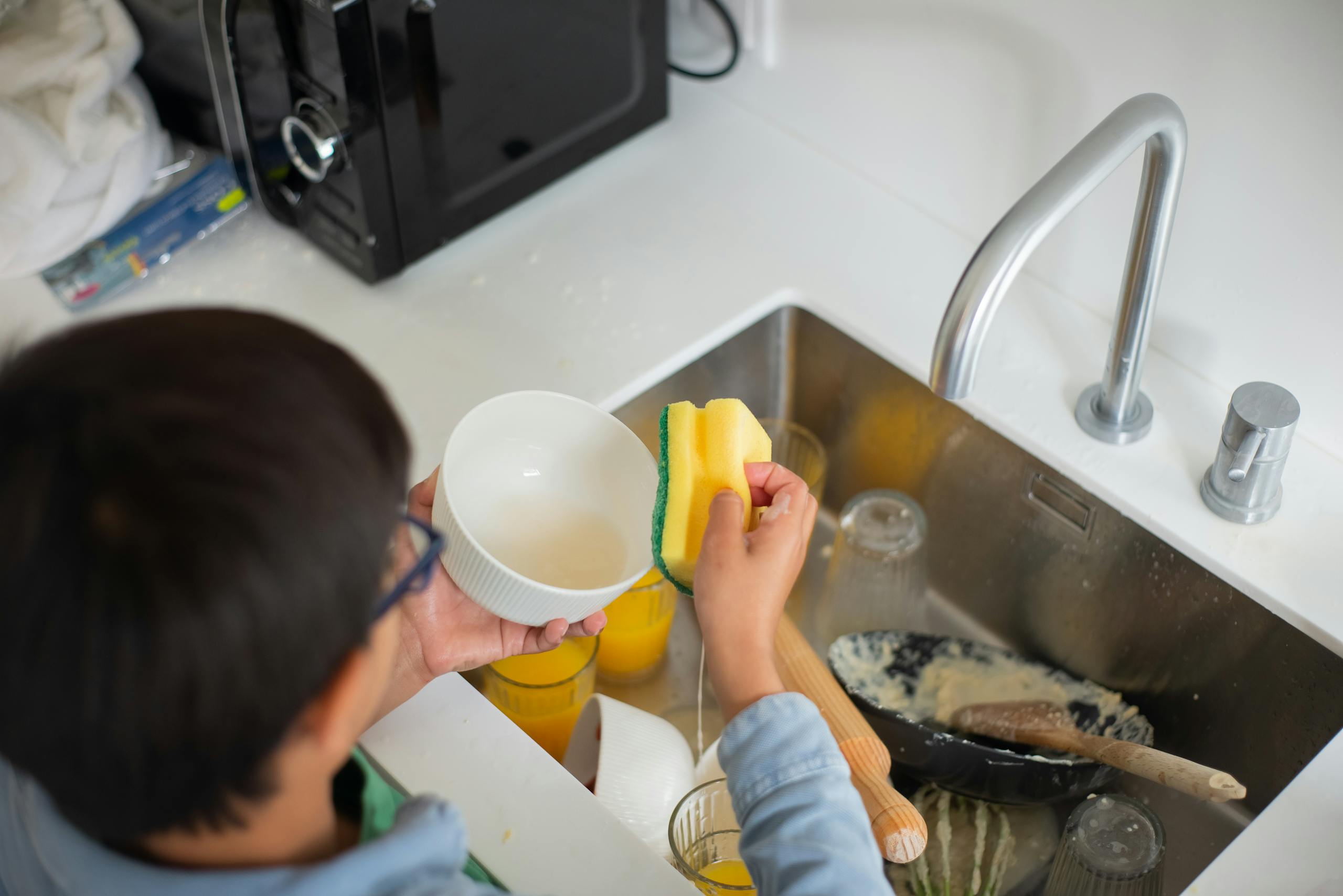 A young boy diligently washing dishes with a sponge in the kitchen sink.